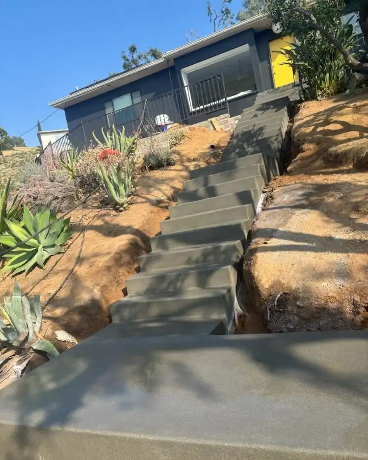 New concrete steps climbing a steep hillside planted with agave toward a dark blue house