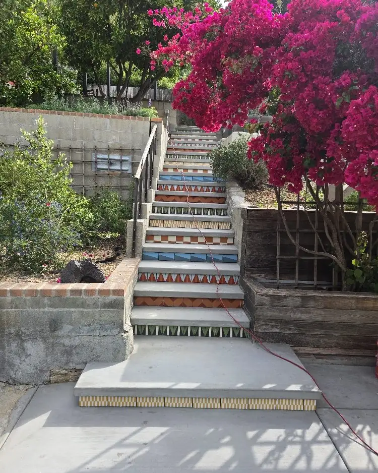 A hillside stair with painted tile risers climbing between block walls under a blooming bougainvillea, with a new concrete landing at the bottom